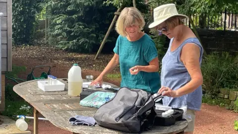 Jane Davis Two women in their sixties stand at a table on which there are various testing apparatus. They are talking together and discussing results 