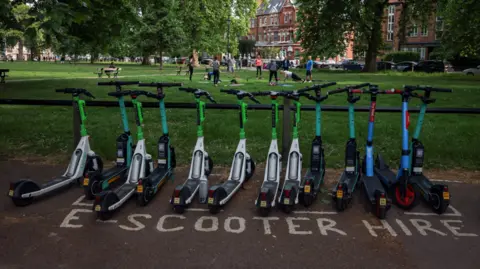 A row of different coloured rented e-scooters lined up in a park in a box demarcated by white lines on the floor and labelled 'E-scooter hire'