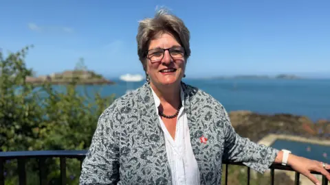 A woman with short grey hair smiling at the camera. She has rectangular glasses on. She is wearing a grey jacket that has a floral pattern and a white blouse underneath. She is wearing a dark, beaded necklace.