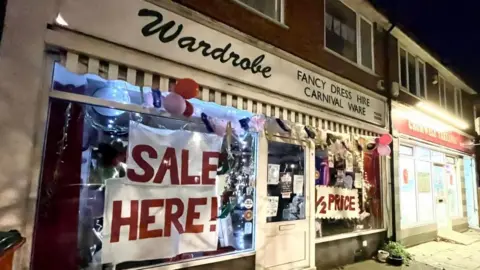 Brian Farmer/BBC The Wardrobe shop front. A sign said "Wardrobe fancy dress hire, carnival ware". Red and pink balloons hang from the shop. Signs written in red capital letters say, "Sale Here" and "1/2 Price" 