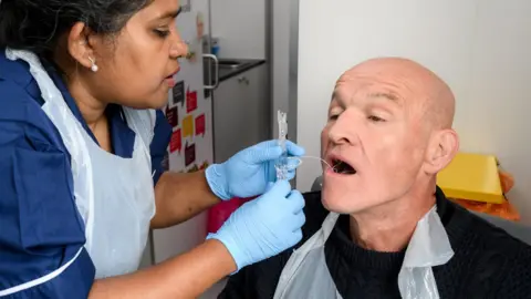 A nurse in blue scrubs wearing gloves pushes a thread into the mouth of a man with a bald head and a black jumper