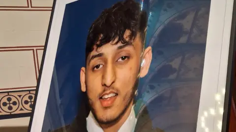 A photo of PC Faizaan Najeeb, pictured in uniform, is printed and framed. It is fixed to an easel in a hall.