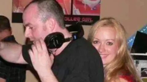 PA Life Tim Burgess, who has a black t-shirt on and a pair of headphones around his neck, is embraced from behind by his sister Linzi Stewart, wo has long blonde hair and wears a red top