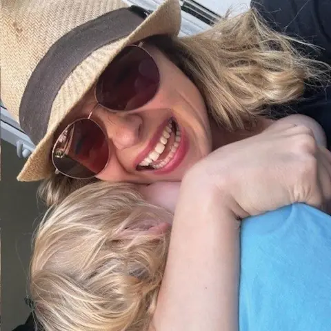 Kimberley Nixon A blonde woman wearing a sunhat, sunglasses and black top. She has a blonde toddler cuddled to her chest, who is wearing a blue t shirt.