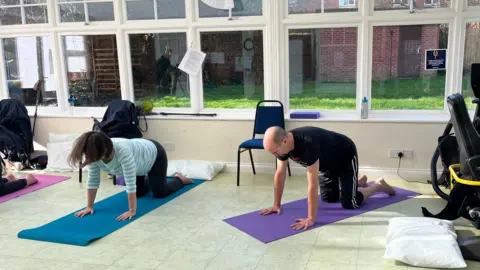 Two people can be seen on yoga mats in a yoga class being held in a conservatory. There is a wheelchair nearby. 