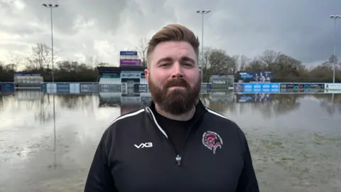James Tolman is standing by a flooded rugby pitch looking at the camera. He is wearing his team's kit which is black with the club's logo in red and white in the corner of his chest