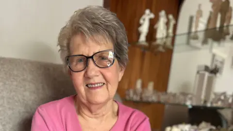 An elderly woman in a pink top with tinted glasses sits smiling in a chair in front of a cabinet of awards and ornaments.