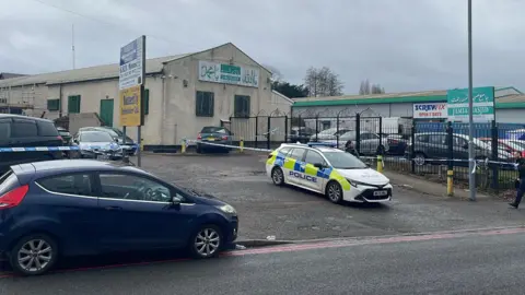 A police car and several other vehicles parked outside a white building with green metal grilles over the windows and a white sign painted in green with indistinguishable English and Arabic text. A sign on the pavement reads Jamia Masjid, along with Arabic text.