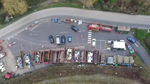 A drone shot of the Lower Compton recycling centre. A row of large metal skips can be seen from above, with parking spaces next to them and several cars parked up. There is a road next to it and greenery round the edges.