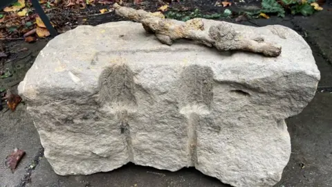 Stuart Whincup/BBC A large block of stone with trowel-shaped markings carved in it sits on top of a concrete surface. It has a knobbly tree branch on top of it. 