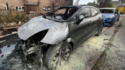 A burnt-out car in front of terraced houses, with its charred engine fully exposed