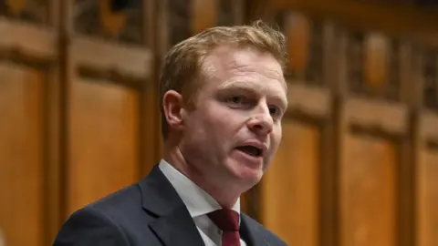 A man with short red hair speaks in the House of Commons. He is wearing a dark coloured suit and a red tie. Behind him is a panelled wall