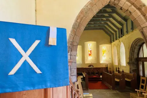 Interior of church building - Arbuthnott Church - with narrow stained glass windows in the background, and a pullpit in the foreground, with a white x on blue material draped over it.