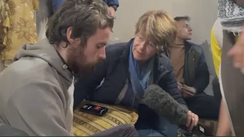 a man sits on a sofa looking forward as a woman holding looks at him 