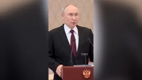 Vladimir Putin has short hair and wears a black suit as he stands at a podium and speaks into a microphone