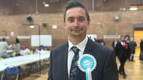 Claire Hamilton/BBC George Woodward, in his late 20s, who has brown hair styled into a quiff, smiles at the camera while wearing a dark grey suit, white shirt and floral tie. He is wearing a Reform rosette. 