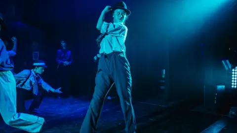 Dave Merritt Erin Rose performing in a theatre show. The stage is lit with blue spotlights and Erin is in the centre of the shot wearing trousers, a shirt and a hat. She is holding the hat and looking to the side. 