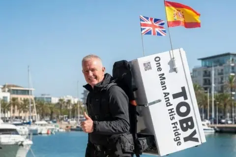 First Contact UK Mental Health Thomas Judge, a man in his 60s with short,grey hair, is standing close to the water's edge on a boardwalk at a marina, apparently in Spain. It's sunny and he is carrying a white fridge on his back with the words "one man and his fridge" and the name Toby (after a baby who died) printed on the side. 