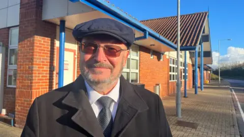 Andrew Turner/BBC Graham Plant, wearing a flat cap and tinted spectacles. He has a greying beard and moustache, and is wearing a white shirt, grey tie, and black coat. He is pictured outside the shelter building at Postwick Park and Ride on the east of Norwich. The building is built of red brick and red concrete tile, with blue pillars, gutters and fascia panels, and white windows and some cladding panels in the gables.