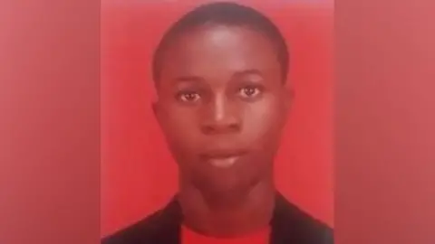 A low-resolution headshot of Segun Olowookere. He has short hair and is wearing a red t-shirt and dark jacket