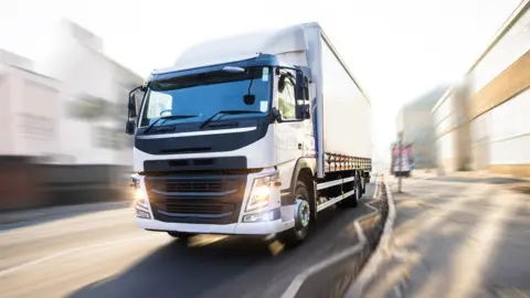 A white lorry driving down an urban street. The lorry has it's lights on. The street is blurred.