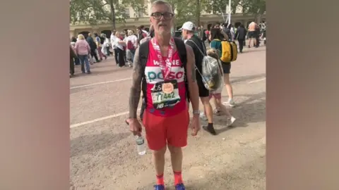 Wayne Goodhead Wayne has grey hair and black glasses and is wearing a pink vest with white writing that reads "Wayne, PDSA". His race bib is black with green numbers on it. He has a medal around his neck which has a pink and white ribbon with a bronze coloured medal