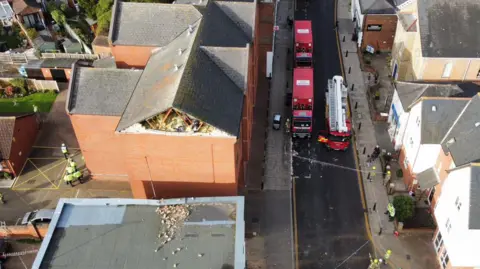 Maldon Fire Station Drone image of a fire engine and two red lorries on an otherwise vehicle-free High Street. Several fire fighters and police officers are standing around. In the centre of the image a red-brick building has its gable end of bricks missing. Rubble can be seen on a neighbouring flat roof at a lower level next to it.