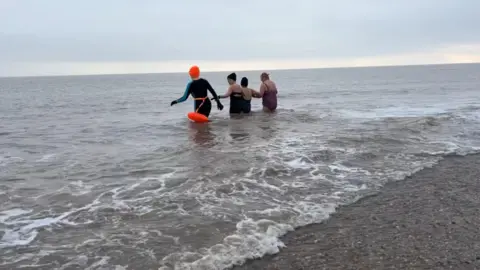 Andrew Turner/BBC Pam Spychal, dressed in an orange hat and black swim suit, and orange floatation marker, with three friends entering the sea at Great Yarmouth beach