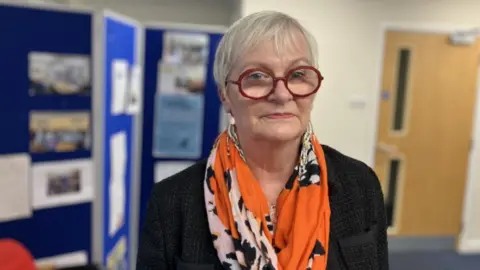 CEO of Headway Gloucestershire Julie Reader-Sullivan is looking at the camera. She is standing in an office-like building; the background is not in focus. She has short hair and is wearing round glasses, a patterned scarf and a dark jacket.
