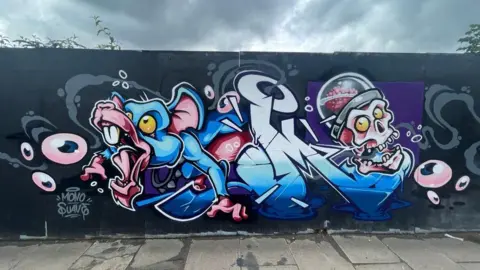 James Thompson/Wall Games A street art mural featuring a blue rat that is escaping the middle of the painting while a skeletons head on the right of the picture seems to be shouting at it. There are eyeballs floating around.