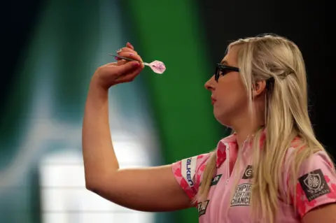 PA Media Fallon Sharrock, a blonde woman in glasses and a pink top, throws a dart