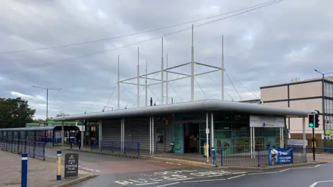 A view of the Bury St Edmunds bus station. A single storey building can be seen with a bus lane running past it to the left. Bus shelters can be seen behind it.