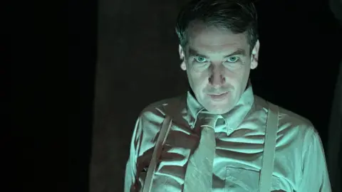 A man is staring at the camera. It is dimly lit and his clothing appears to be green