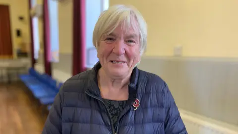 A smiling grey-haired woman in a blue jacket with a poppy badge on it stands in a village hall