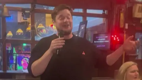 Man in black sweater closes his eyes as he sings into microphone - a woman in a white shirt with blonde hair sits behind him. You can see the words on karaoke machine.