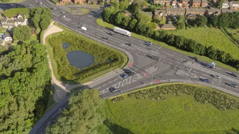 CGI view of a t-junction, showing traffic on two roads converging surrounded by green fields and trees.