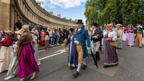 Matt Cardy/Getty Images A large crowd of people walking in a parade through Bath, wearing full Regency attire and carrying props like parasols, fans, baskets and top hats. 