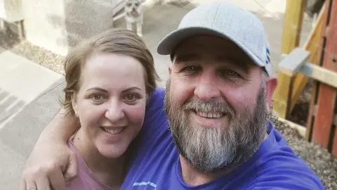 Martin Sutherland A man with a longer beard is standing in front of a house with his arm around a woman with short blond hair. She is wearing a pink top, while he is dressed in a dark-blue t-shirt and a grey cap. There is a schnauzer dog visible behind the couple.