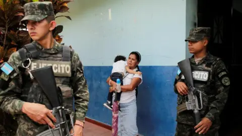 REUTERS/Leonel Estrada Membros das forças de segurança montam guarda enquanto uma mulher carrega sua filha enquanto espera do lado de fora de uma seção eleitoral, enquanto a contagem continua em San Antonio de Flores, Honduras, em 7 de dezembro de 2025.