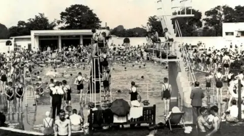 The Bournville Society A black and white photo of Rowheath Lido. The rectangular pool is packed full of people in the water and on the outside. Several people queue up for a slide that is about 10 metres tall. Next to it three to four people stand on a diving board. 