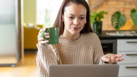 A young woman holds a green mug and looks exasperated at an open laptop