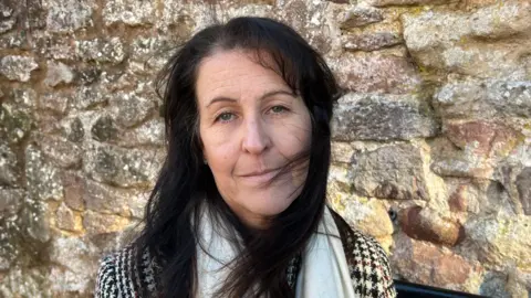 Siobhan, a woman with long dark hair, looks at the camera, standing in front of a stone wall. She is wearing a white scarf and dogtooth-pattern coat.