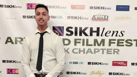 Kay S. Ubhi A man in a white shirt and black tie stands in front of a white banner which says Sikh lens on it, aslongside other icons.