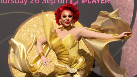 BBC/World Of Wonder /Kieron McCarron La Voix is wearing a large gold dress with a large glittery circular back at a launch event for RuPaul's Drag Race UK.