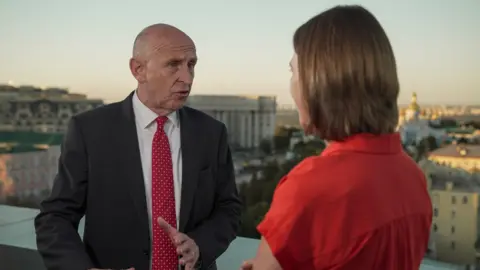BBC John Healey, wearing a dark jacket, white shirt and red dotted tie facing Sarah Rainsford wearing a red top