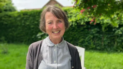 Lesley Clarke successful  her plot  connected  a sunny day. She is smiling into the camera and wearing a blouse with bladed  vertical stripes and a grey cardigan. The writer  and precocious   hedge successful  the inheritance  are blurred.