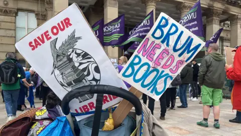 BBC Save our libraries signs in a bag, with protesters in the background demonstrating against cuts to services in Birmingham