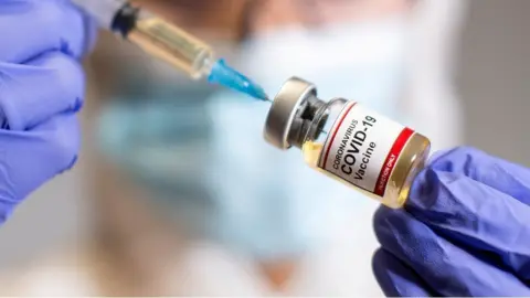 A needle going into a phial of Covid-19 vaccine. It is being held by someone wearing blue medical gloves