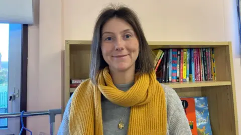 Lucy Hoyle stands in front of a wooden bookshelf filled with brightly-coloured children's books. She wears a grey jumper, a yellow scarf and a long gold necklace. She has short brown hair.