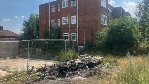 The piled-up remains of a burnt-out caravan on Baguley Crescent. The image shows debris next to metal fencing and scrub land in front of an abandoned block of flats.